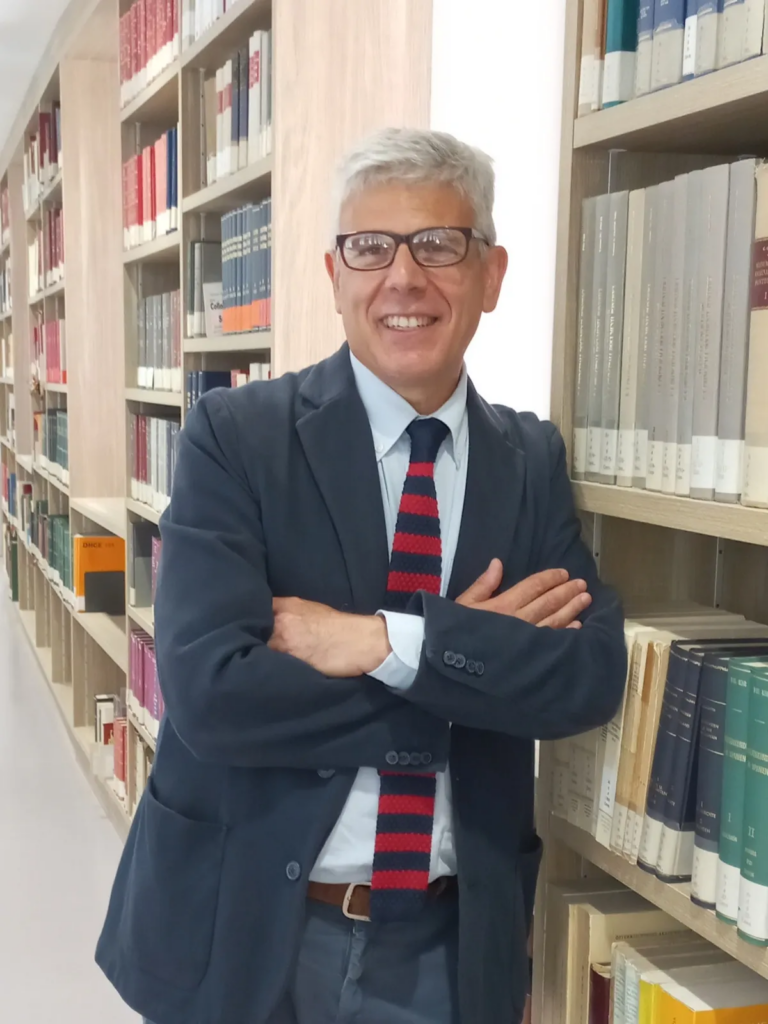 Portrait de l'écrivain Giuseppe Lupo, souriant et les bras croisés, posant devant les rayonnages d'une bibliothèque. Il porte des lunettes, une veste de costume sombre et une cravate à rayures horizontales rouges et bleues.